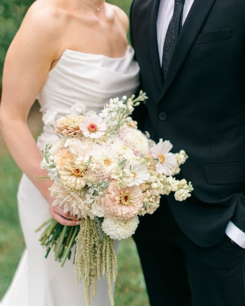 Bride holding a peach and cream bouquet with trailing amaranthus, standing beside the groom