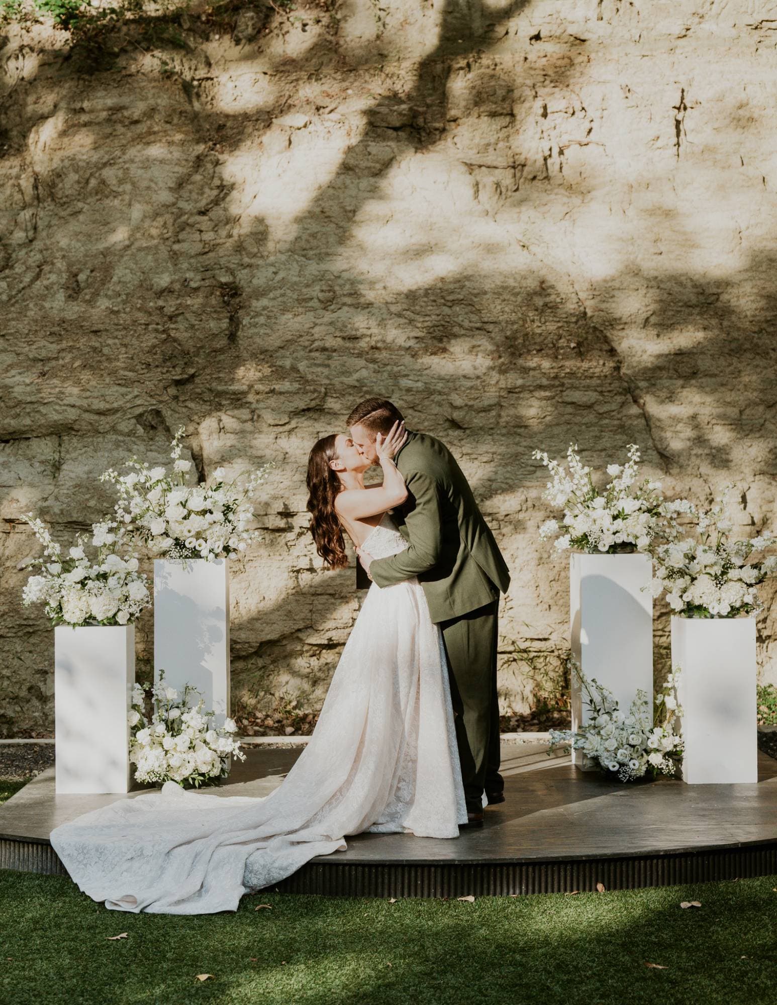 Bride and groom sharing a kiss at their ceremony, flanked by white floral arrangements